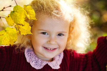Fototapeta premium A child in an autumn park flooded with the sun. A little blonde girl in a bard knitted sweater among yellow leaves