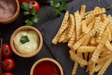Homemade baked potato fries with mayonnaise, tomato sauce and rosemary on paper over black stone background. . tasty french fries, unhealthy food.