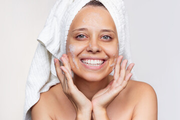 A young beautiful caucasian smiling woman with a white towel on her head after a shower cleanses her face with foam for washing on a white background. Skin care, cosmetology. Morning routine. Close up