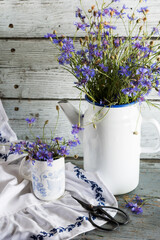 Bouquet of just cut cornflowers  in a metal  teapot and in cup, scissors on white tablecloth on wooden background, floral still life concept