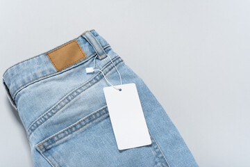 Jeans clothing white paper tag, label blank mockup template, to place your design. On a blue jeans and grey background, copy space, flat lay