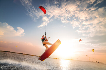 Sports shot of a young caucasian woman in a wetsuit doing a trick in the air against the backdrop of a sunset in the sea. Kitesurring girl athlete flies on a kite with a board - obrazy, fototapety, plakaty