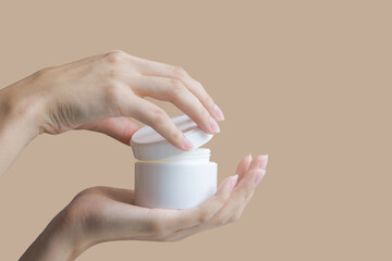 Women's hands open mock-up of white cosmetic package. Two hands hold round jar of body cream on beige background. Concept of beauty industry.