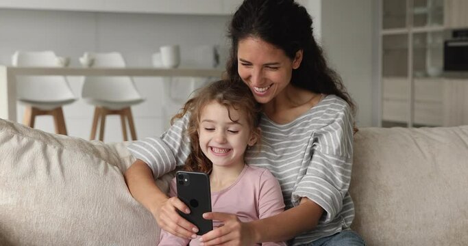 Joyful young nanny playing mobile games with adorable small child girl, resting on comfortable sofa in living room. Addicted to modern technology happy mum and kid daughter having fun using cellphone.