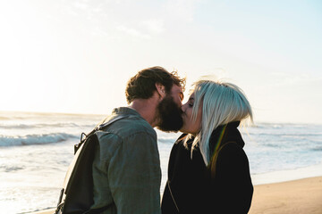 Fototapeta premium Romantic happy couple in love kissing on shoreline at the wild beach - Boyfriend and girlfriend wearing cool clothing and vintage backpack standing in front of the sea during the sunset