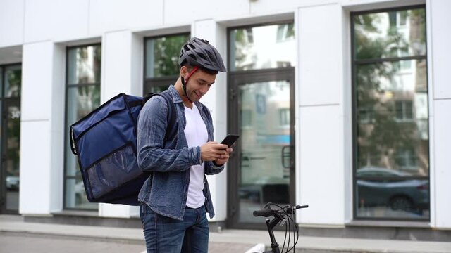 Cheerful handsome male courier with thermo backpack standing with bicycle in city street and using navigation app on mobile phone. Delivery man looking for client address using smartphone, slow motion