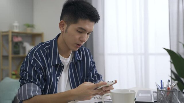 portrait attractive asian guy is mumbling while texting phone message with concentration at the office desk in a bright living room at home.