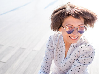 Wind ruffles short hair of freckled woman in colorful sunglasses. Smiling woman at open wooden...