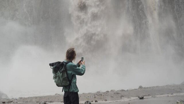 Karakaya-Su, Russia – July  25 2021: 4K Man traveler with backpack walking and watching, taking pictures on cellphone camera of big waterfall in Jilly Su (Dzhily-Su tract) in Elbrus region, Caucasus.