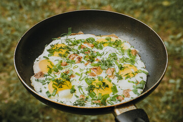 Frying pan of fried eggs, bacon and organic greens, green grass on the background, cooked on the grill on a summer sunny day, summer breakfast, eating in nature - the concept of a healthy lifestyle