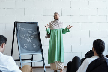 Young female speaker in hijab standing by blackboard in front of audience