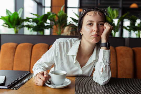 Sad and tired female entrepreneur leaning on hand while sitting at table in cafe during coffee break and looking away 