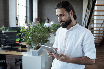 Young Arabic businessman in white shirt looking at tablet screen while scrolling through online data