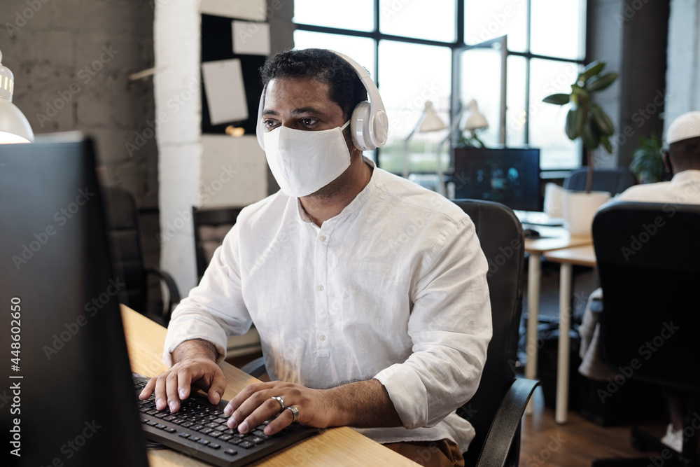 Poster serious mixed-race businessman in mask and headphones looking at computer screen while typing