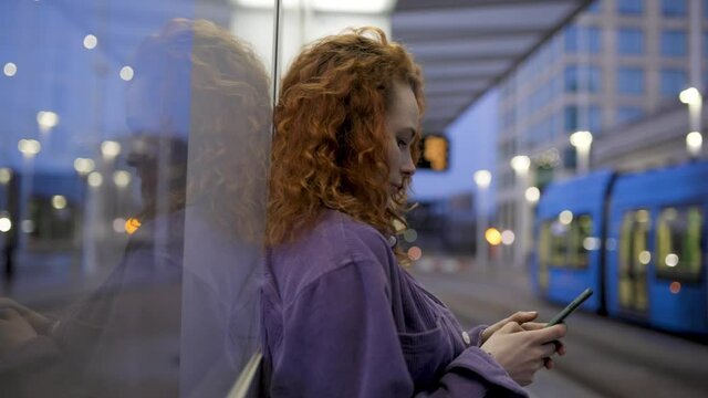 Pretty woman leaning on glass pane at train station using smartphone