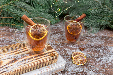 Two glass cups of tea with lemon, cinnamon sticks on a wooden table against the background of a green Christmas tree with lights garlands. New Year Christmas card.
