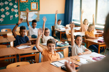 Group of happy elementary students raising their hands to answer teacher's question during class at school.