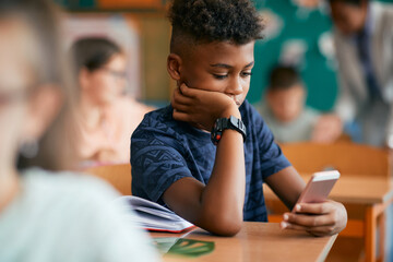 Black elementary student typing text message on smart phone in classroom.