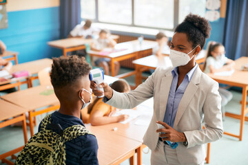 Black teacher checking temperature of schoolboy while he's entering classroom due to COVID-19 pandemic.