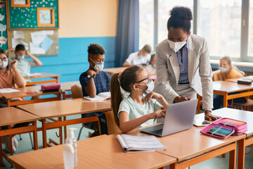 Elementary student and her teacher wearing face masks while using laptop during class at school.