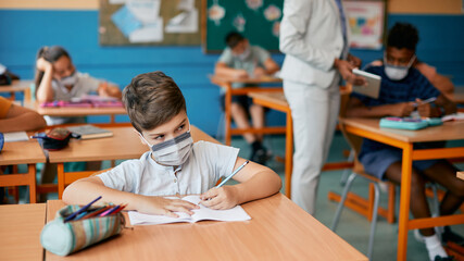 Schoolboy with protective face mask writing while having class in classroom during coronavirus pandemic.