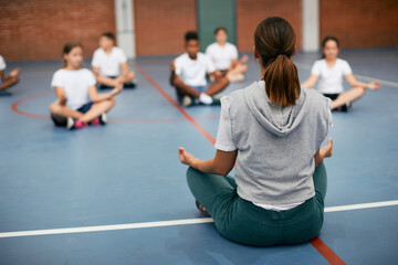Rear view of sports teacher practicing Yoga with her students at school gym.
