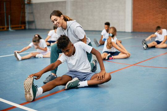 Happy female sports teacher assisting African American boy with stretching exercise at school gym.