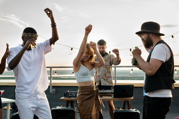 Young intercultural friends and happy deejay dancing together at rooftop party