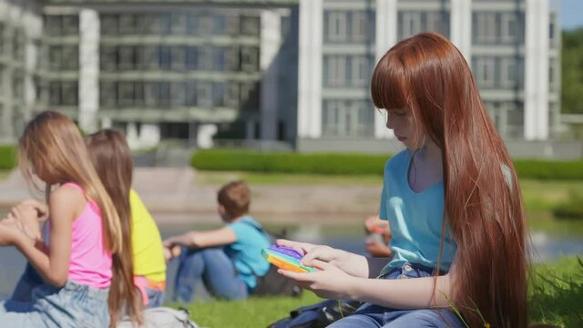 Preteen caucasian schoolgirl sitting on grass outdoors playing pop it toy