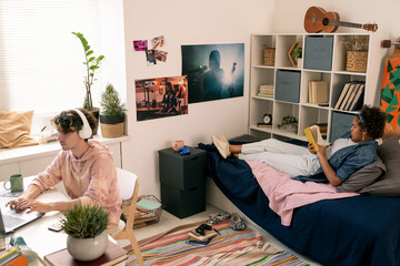 Two contemporary restful teenagers reading book and networking in bedroom