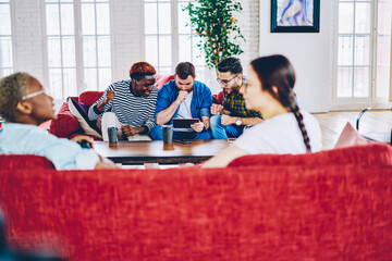 Tense multiracial male friends browsing website on digital tablet searching information during free time, emotional men watching streams video of match using touchpad device in loft apartment