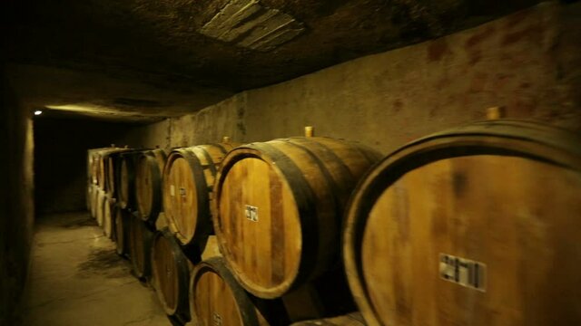 Aged wooden barrels with a wine , brandy or whiskey in a big warehouse . Dolly , ronin movement a long row of wine barrels at a vintage winery with alcohol . 