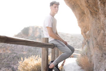 Chico joven rubio reflexivo sentado haciendo senderismo junto a las montañas en la puesta de sol