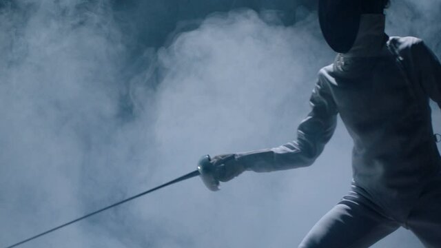 Professional fencer in protective uniform practicing maneuvers with sword alone in dark studio . Fencing demonstration on black smoked background . Shot on Arri Alexa cinema camera in slow motion .