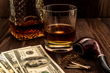 Glass of whiskey with a money and tobacco pipe with tobacco leaves are scattered on the wooden table. Angle view