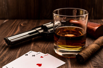 Glass of whiskey and playing cards with revolver and cuban cigar on the wooden table. Angle view
