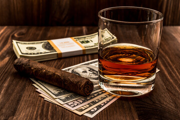 Glass of whiskey and a pack of dollars with cuban cigar on a wooden table. Angle view