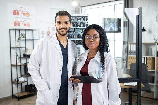 Multiethnic doctors at modern office indoors. Pretty afro-american female doctor holding digital tablet in hands, posing to camera with smile together with male arabian colleague radiologist