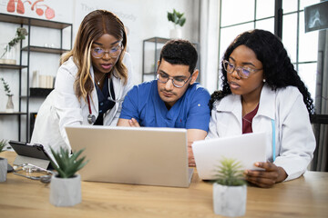 Three focused multiethnic doctors having online conference at hospital boardroom. Male and female heath care workers using modern laptop during meeting.