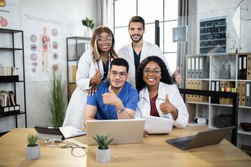 Fototapeta premium Multiracial group of doctors wearing coats and uniforms, sitting together at table and smiling on camera with thumbs up. Medical specialists during session at hospital. Cooperation concept.