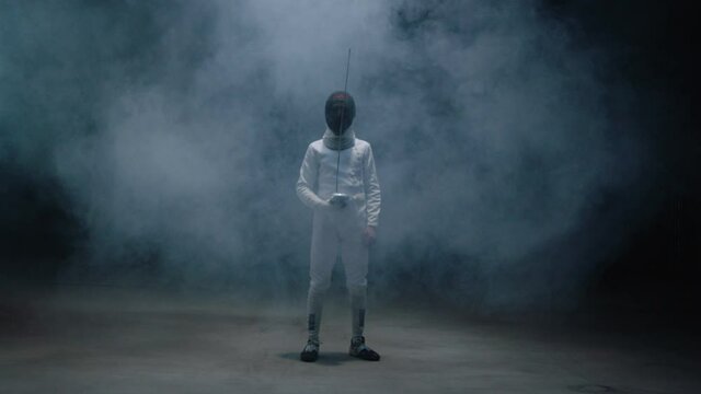 Professional fencer in protective uniform practicing maneuvers with sword alone in dark studio . Fencing demonstration on black smoked background . Fencer stannding and looking to camera . Portrait 