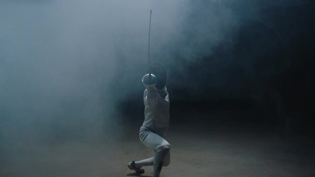 Professional fencer in protective uniform practicing maneuvers with sword alone in dark studio . Fencing demonstration on black smoked background . Shot on Arri Alexa cinema camera in slow motion .