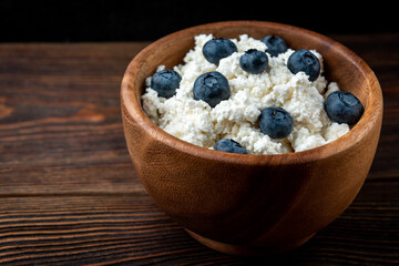 Cottage cheese and blueberry in wooden bowl and spoon on dark wooden background.