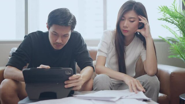 asian marry couple checking and calculate financial billing together on sofa involved in financial paperwork, paying taxes online using e-banking laptop at living room home background