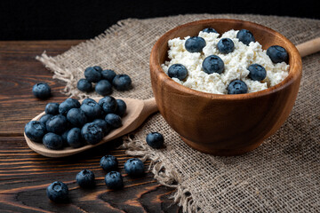 Cottage cheese and blueberry in wooden bowl and spoon on dark wooden background.