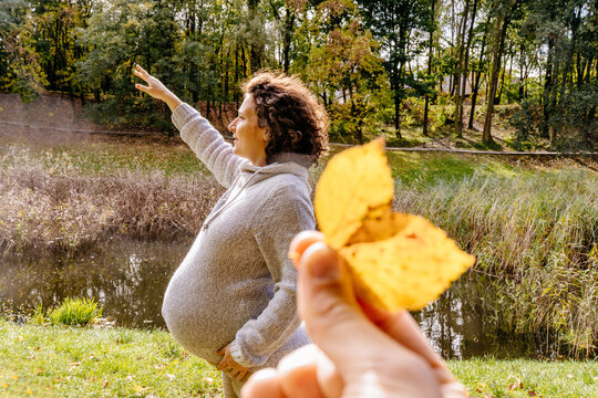 Cozy autumn photo of curly pregnant woman at autumn park. Husbands hand holding leaves like butterfly wings. Towards the dream, future parenting concept.