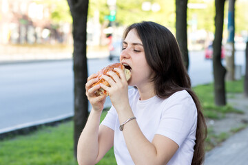 Brunette woman eating burger on the street near the road in the city. Fast food on the way to work or school.