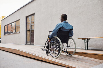 Rear view of handicapped African-American man in casual shirt using wheelchair ramp built by his...