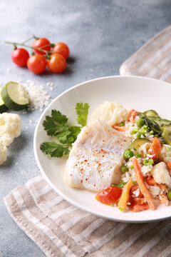 Seafood dish. White fish with rice and vegetables: zucchini, cauliflower, carrots, tomatoes and herbs in a white plate on a gray background. Background image, copy space