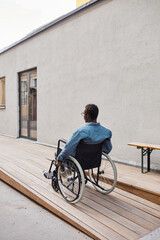 Fototapeta na wymiar Rear view of disabled young African-American man sitting in wheelchair and moving along ramp near house